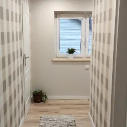 Wąski korytarz z tapetą w beżowo-szarą kratę, drewnianą podłogą i dywanikiem. Widoczne drzwi, okno z rośliną i oświetlenie sufitowe.