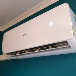 Minifix Michał Kaczmarek - Biała klimatyzacja AUX zamontowana na turkusowej ścianie, widoczny wyświetlacz z temperaturą 16 stopni, rura odprowadzająca skropliny.