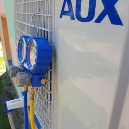 Minifix Michał Kaczmarek - Podłączenie manometrów serwisowych do klimatyzatora marki AUX podczas przeglądu, widoczne węże w kolorach niebieskim i żółtym oraz logo producenta na obudowie.