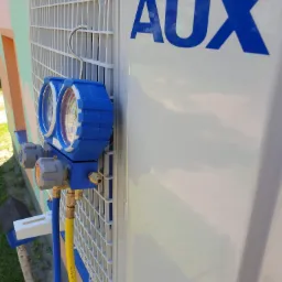 Podłączenie manometrów serwisowych do klimatyzatora marki AUX podczas przeglądu, widoczne węże w kolorach niebieskim i żółtym oraz logo producenta na obudowie.