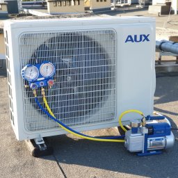 Minifix Michał Kaczmarek - Biała jednostka zewnętrzna klimatyzacji marki AUX na dachu, podłączona do pompy próżniowej VALUE za pomocą niebiesko-żółtych przewodów manometrycznych. Widoczne manometry wskazują ciśnienie.