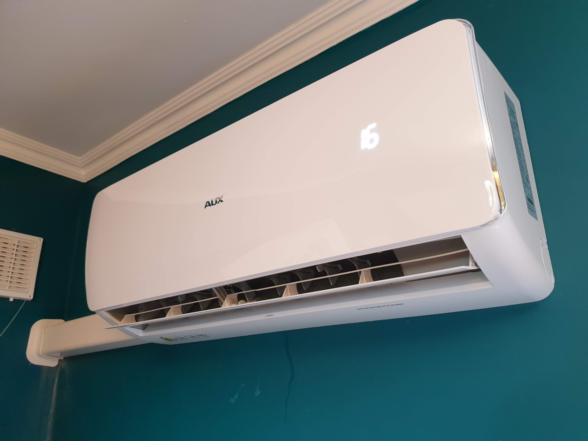 Biała klimatyzacja AUX zamontowana na turkusowej ścianie, widoczny wyświetlacz z temperaturą 16 stopni, rura odprowadzająca skropliny.
