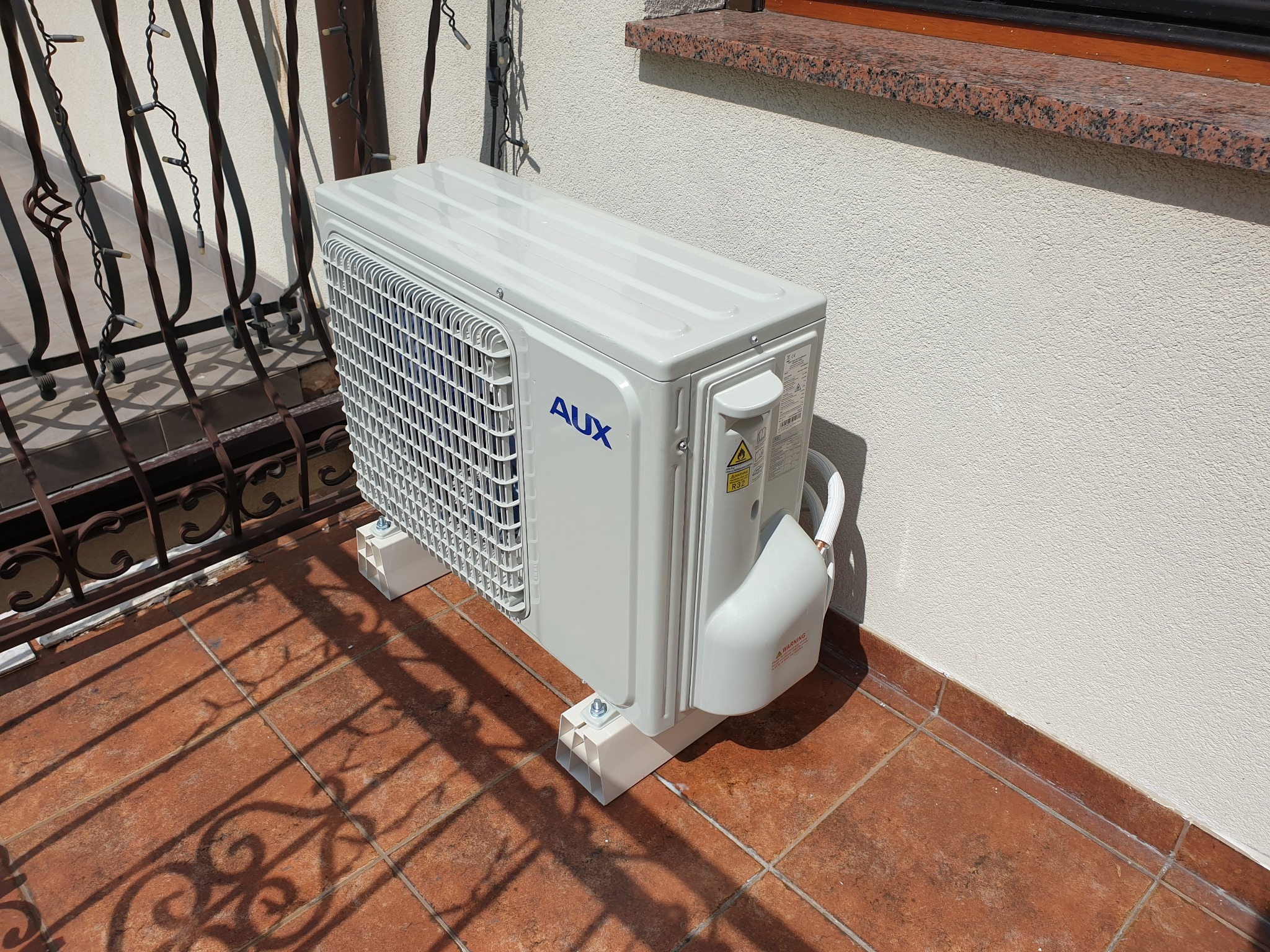 Zewnętrzna jednostka klimatyzacji marki AUX zamontowana na balkonie z terakotową podłogą i ozdobną, metalową balustradą, widoczna etykieta ostrzegawcza.