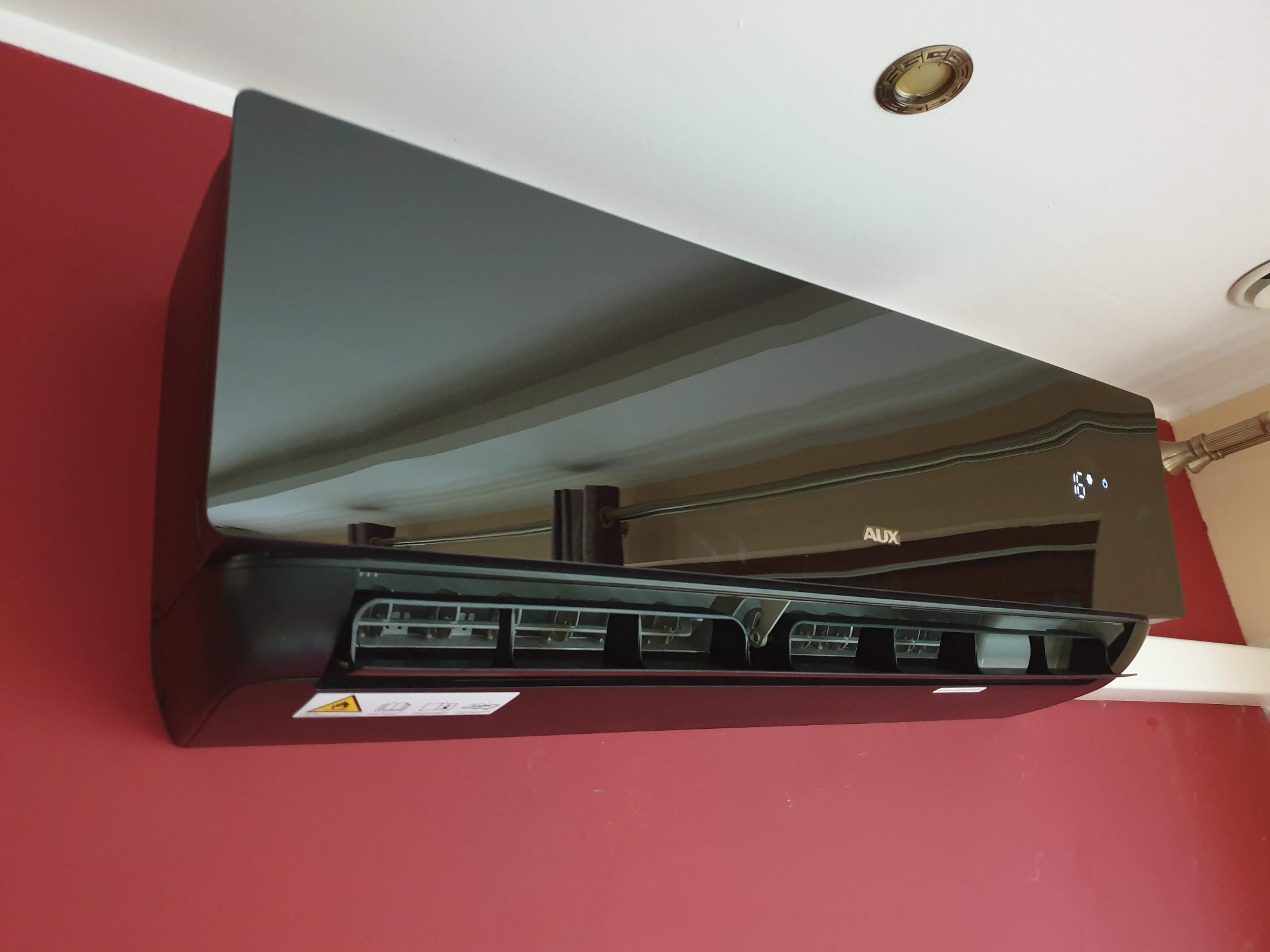 Czarny klimatyzator marki AUX zamontowany na czerwonej ścianie, widoczne odbicie sufitu z oświetleniem w błyszczącej obudowie.