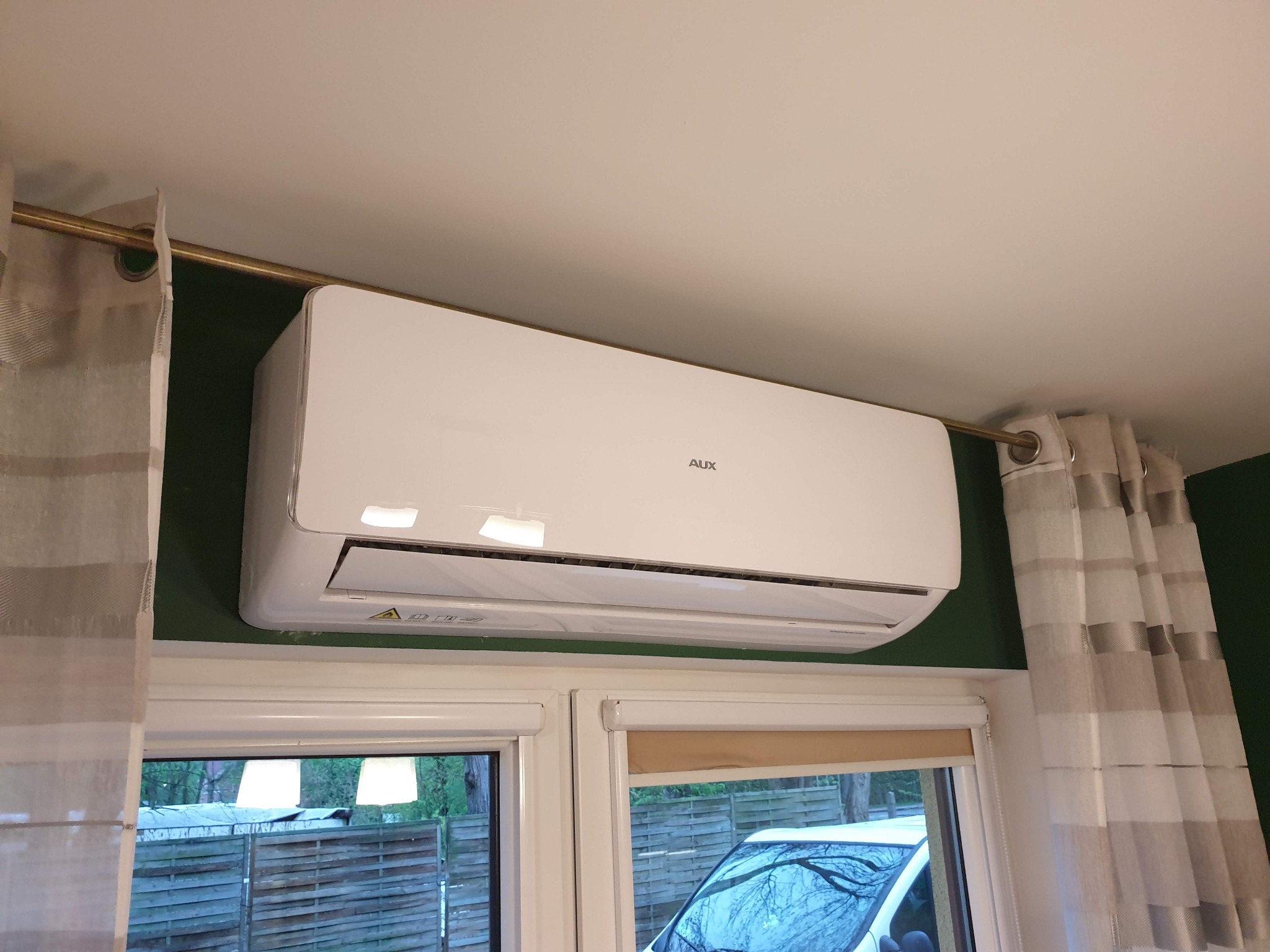 Biała jednostka klimatyzacji AUX zamontowana nad oknem z roletami i zasłonami w pokoju z zielonymi ścianami.
