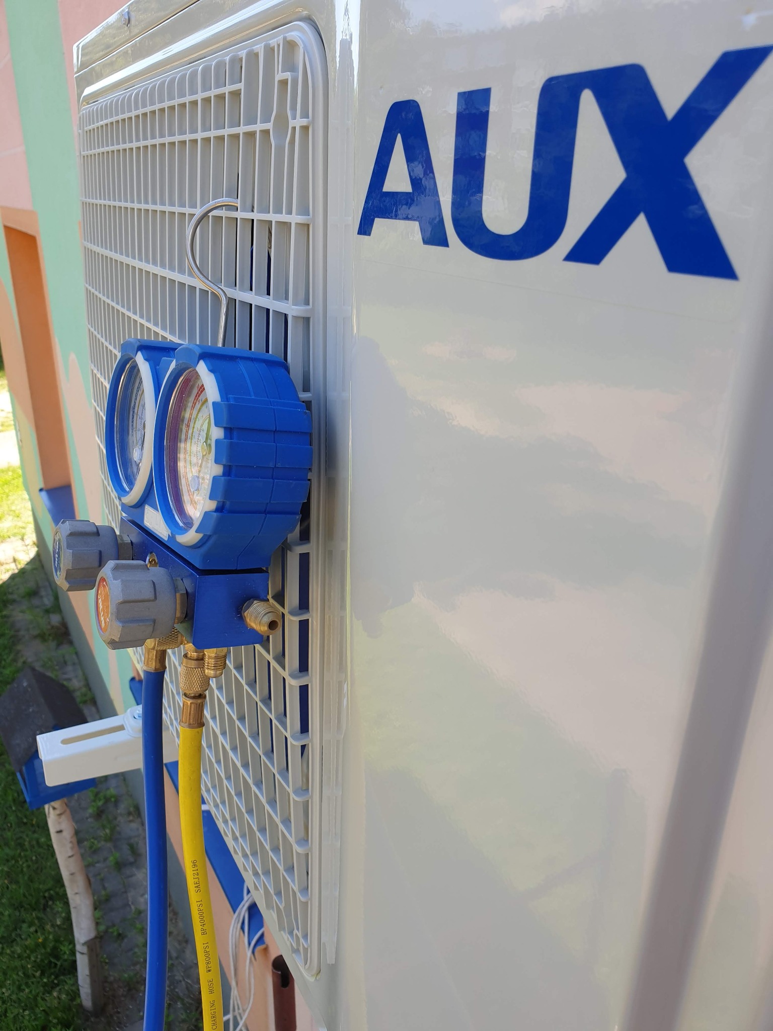 Podłączenie manometrów serwisowych do klimatyzatora marki AUX podczas przeglądu, widoczne węże w kolorach niebieskim i żółtym oraz logo producenta na obudowie.