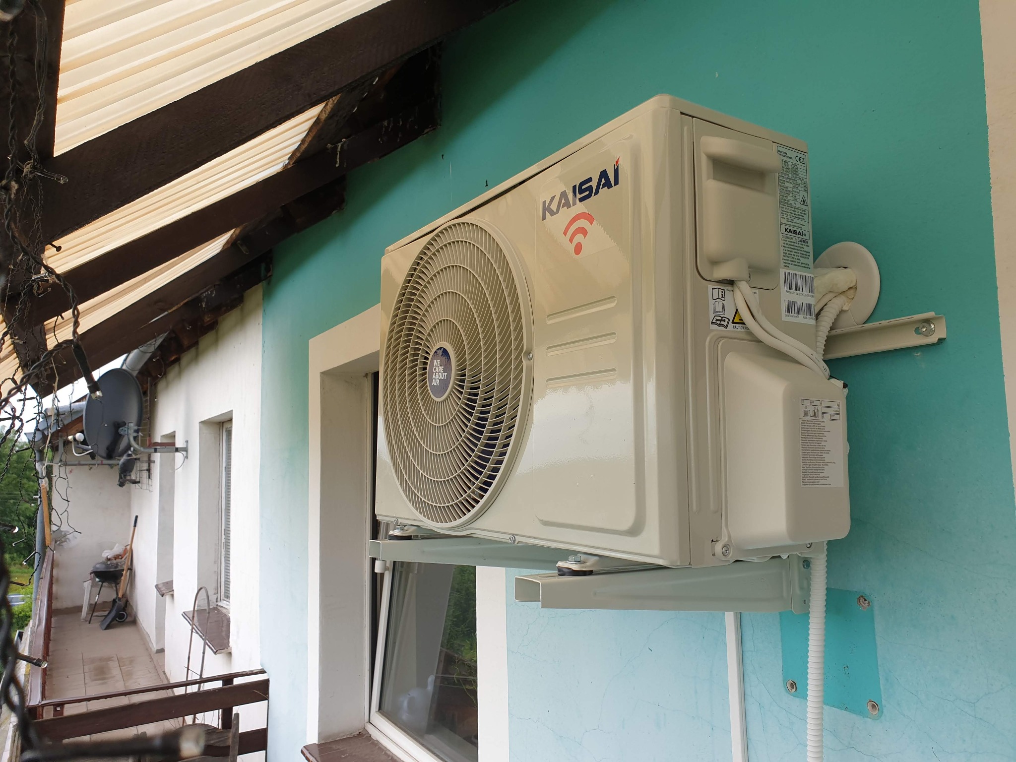 Zamontowana jednostka zewnętrzna klimatyzacji Kaisai na balkonie, widoczne przewody i elementy mocujące na turkusowej ścianie.