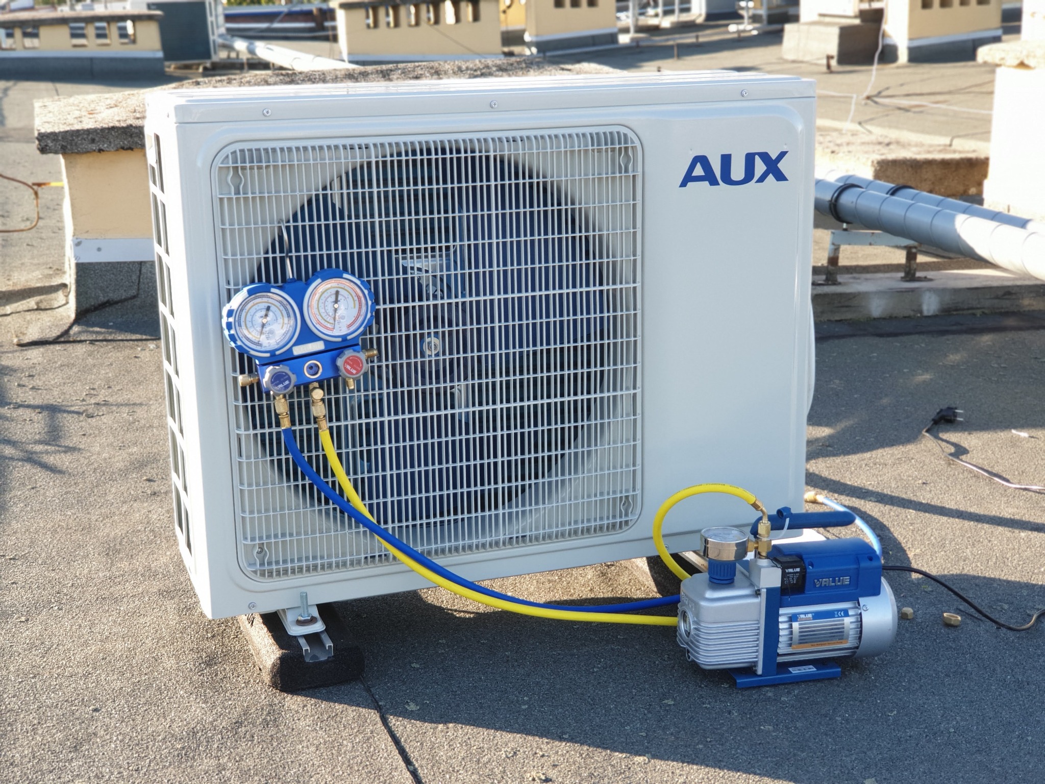 Biała jednostka zewnętrzna klimatyzacji marki AUX na dachu, podłączona do pompy próżniowej VALUE za pomocą niebiesko-żółtych przewodów manometrycznych. Widoczne manometry wskazują ciśnienie.