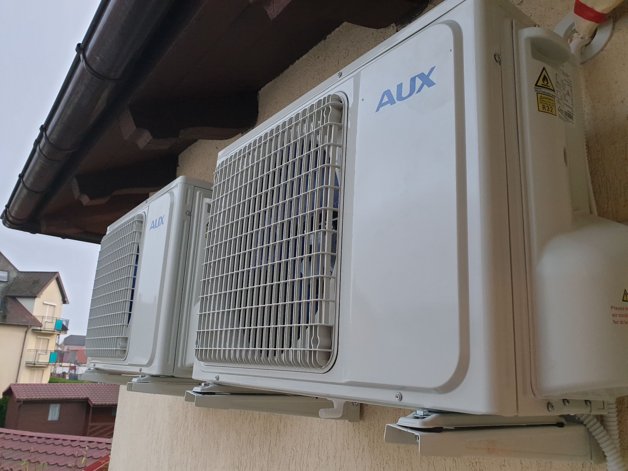 Dwa identyczne, białe klimatyzatory marki AUX zamontowane na ścianie budynku z beżową elewacją, widoczne z zewnątrz, z brązowym dachem i rynną nad nimi, w tle zabudowa mieszkalna.