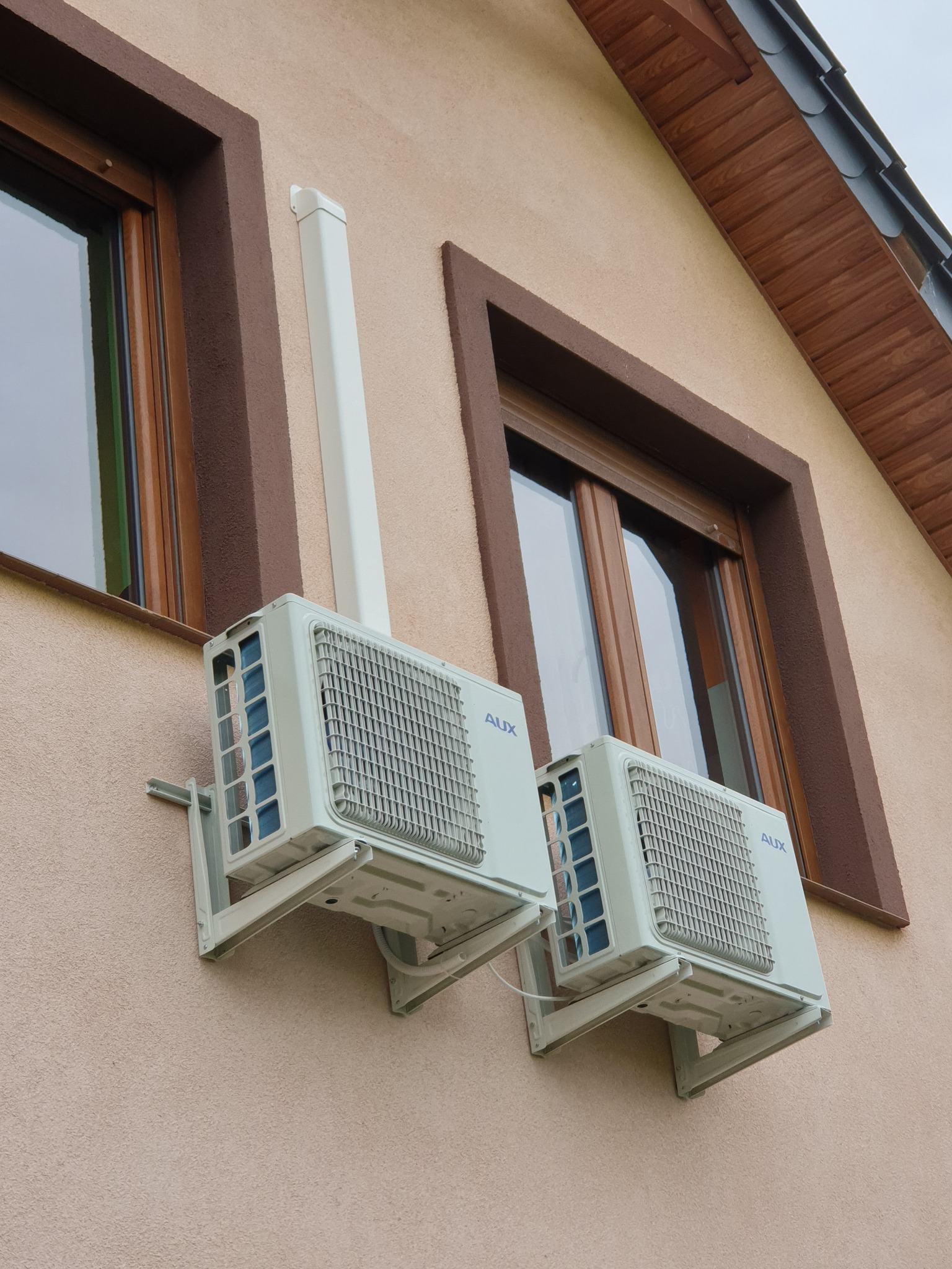 Dwa klimatyzatory marki AUX zamontowane na elewacji budynku z oknami w brązowych ramach. Biała rura odprowadzająca skropliny od jednego z urządzeń.