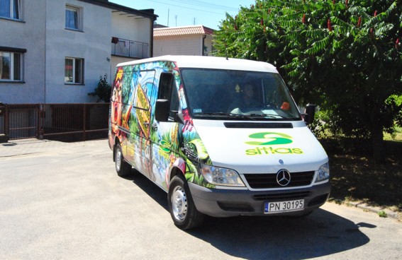 Mercedes Sprinter z kolorową grafiką reklamową na karoserii, logo firmy Sitlas na masce, zaparkowany przed budynkiem mieszkalnym i zielonym drzewem.