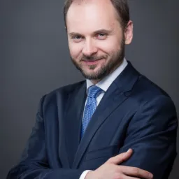 Portret mężczyzny z brodą w granatowym garniturze i niebieskim krawacie, z założonymi rękami i obrączką na palcu, uśmiechający się do kamery na szarym tle.