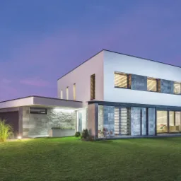 Nowoczesny dom jednorodzinny z szarymi żaluzjami fasadowymi, oświetlonym trawnikiem i garażem, sfotografowany o zmierzchu.