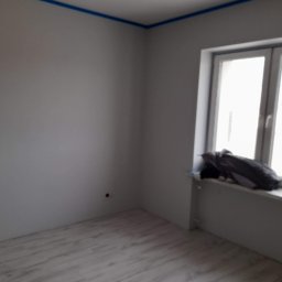 Pająkteam - Pokój po malowaniu ścian na szaro, zabezpieczony niebieską taśmą malarską przy suficie, z widoczną podłogą imitującą drewno i oknem z ubraniami na parapecie.