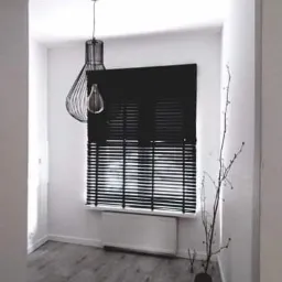 Wnętrze pokoju z szarymi panelami podłogowymi, białymi ścianami, czarnymi żaluzjami w oknie, dekoracyjną lampą wiszącą i gałązkami w wazonie.