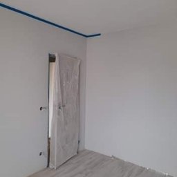 Pająkteam - Pokój po świeżym pomalowaniu na szaro, z zabezpieczoną folią drzwiową i niebieską taśmą malarską przy suficie, gotowy do dalszych prac wykończeniowych.