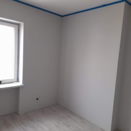 Pająkteam - Pusty pokój z oknem, świeżo pomalowanymi szarymi ścianami i drewnianą podłogą, zabezpieczony niebieską taśmą malarską przy suficie.