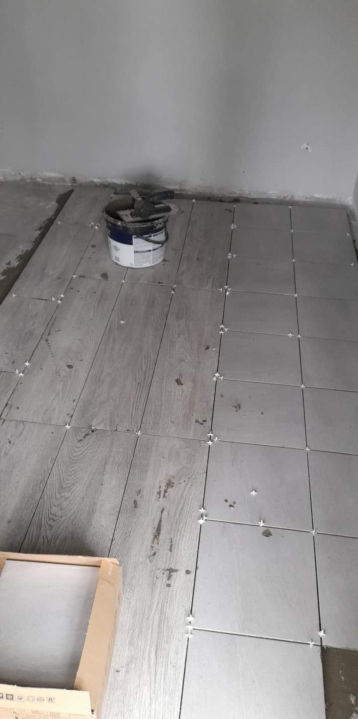 Układanie szarych płytek imitujących drewno, z białymi krzyżykami dystansowymi, widoczny pojemnik z klejem i paczka płytek w rogu