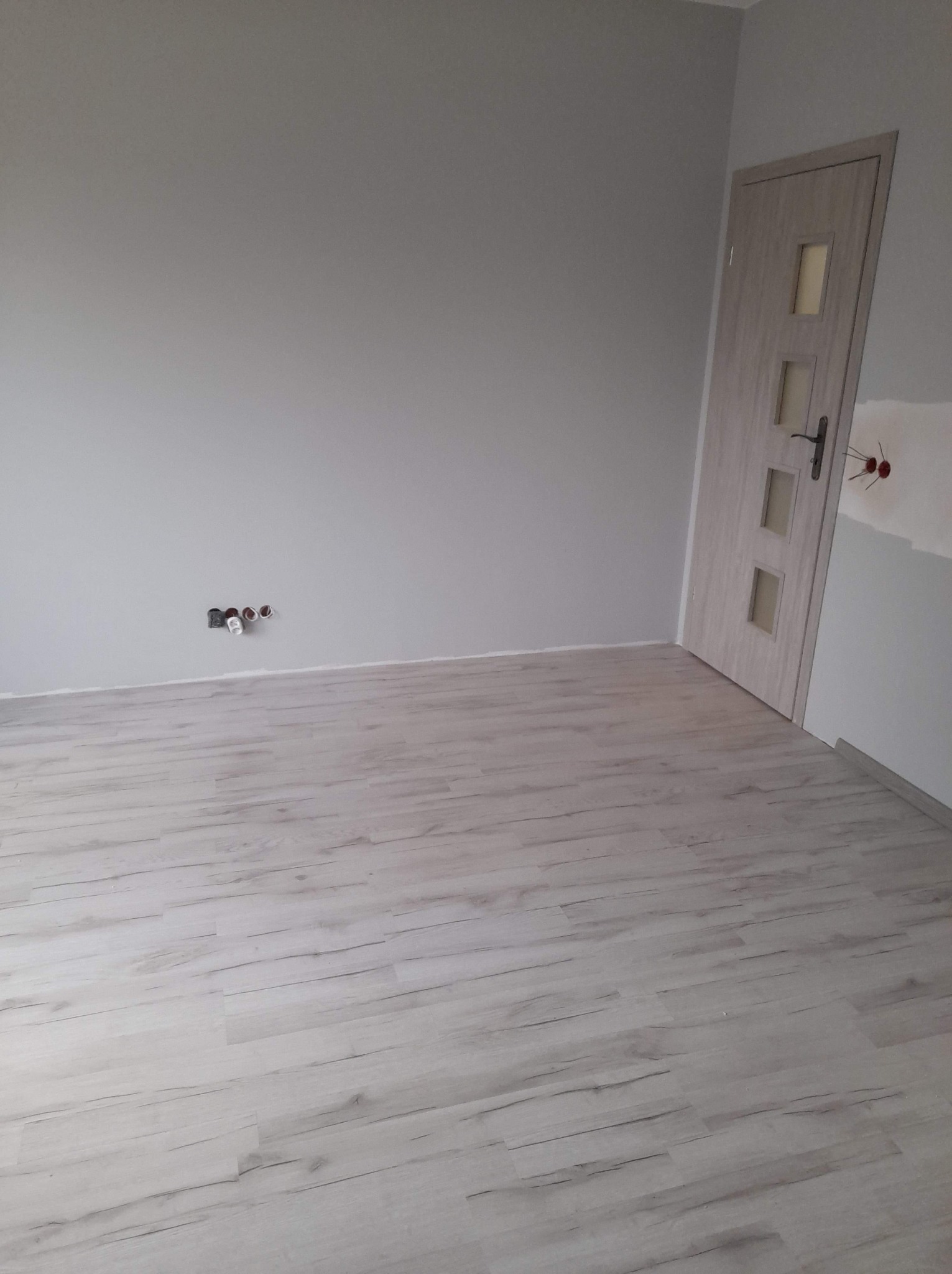 Świeżo pomalowane na szaro wnętrze pokoju z panelami podłogowymi, widoczne gniazdka elektryczne bez osprzętu i fragment ściany z widoczną warstwą szpachli przy futrynie drzwi.