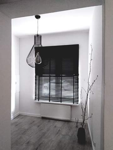 Wnętrze pokoju z szarymi panelami podłogowymi, białymi ścianami, czarnymi żaluzjami w oknie, dekoracyjną lampą wiszącą i gałązkami w wazonie.