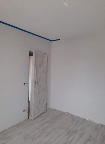 Pokój po świeżym pomalowaniu na szaro, z zabezpieczoną folią drzwiową i niebieską taśmą malarską przy suficie, gotowy do dalszych prac wykończeniowych.