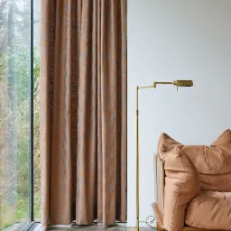 Zasłony w odcieniach brązu i szarości, wiszące przy oknie z widokiem na zieleń, obok lampy podłogowej w kolorze złotym i fragmentu sofy w odcieniu beżowym.