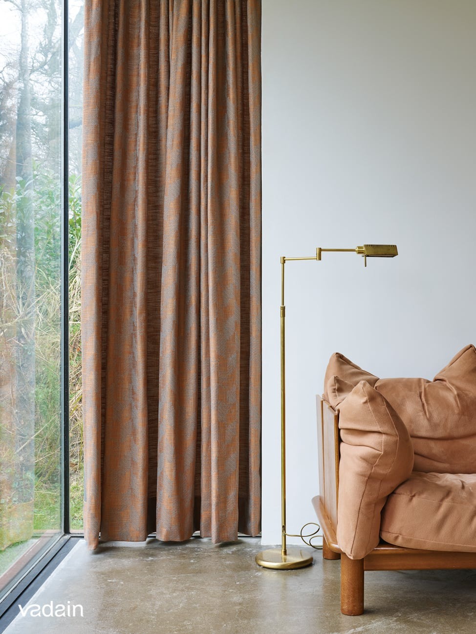 Zasłony w odcieniach brązu i szarości, wiszące przy oknie z widokiem na zieleń, obok lampy podłogowej w kolorze złotym i fragmentu sofy w odcieniu beżowym.