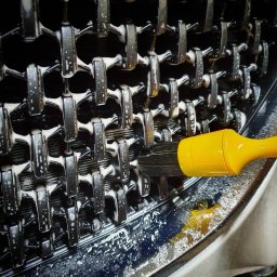 DA Studio Mobilny Auto Detailing - Szczoteczka z żółtą rączką czyści chromowany grill samochodu pokryty pianą.