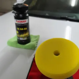 Butelka pasty polerskiej Sonax Profiline EX 04-06 na zielonej ściereczce, obok żółta gąbka polerska na czerwonej ściereczce, na tle białego samochodu.