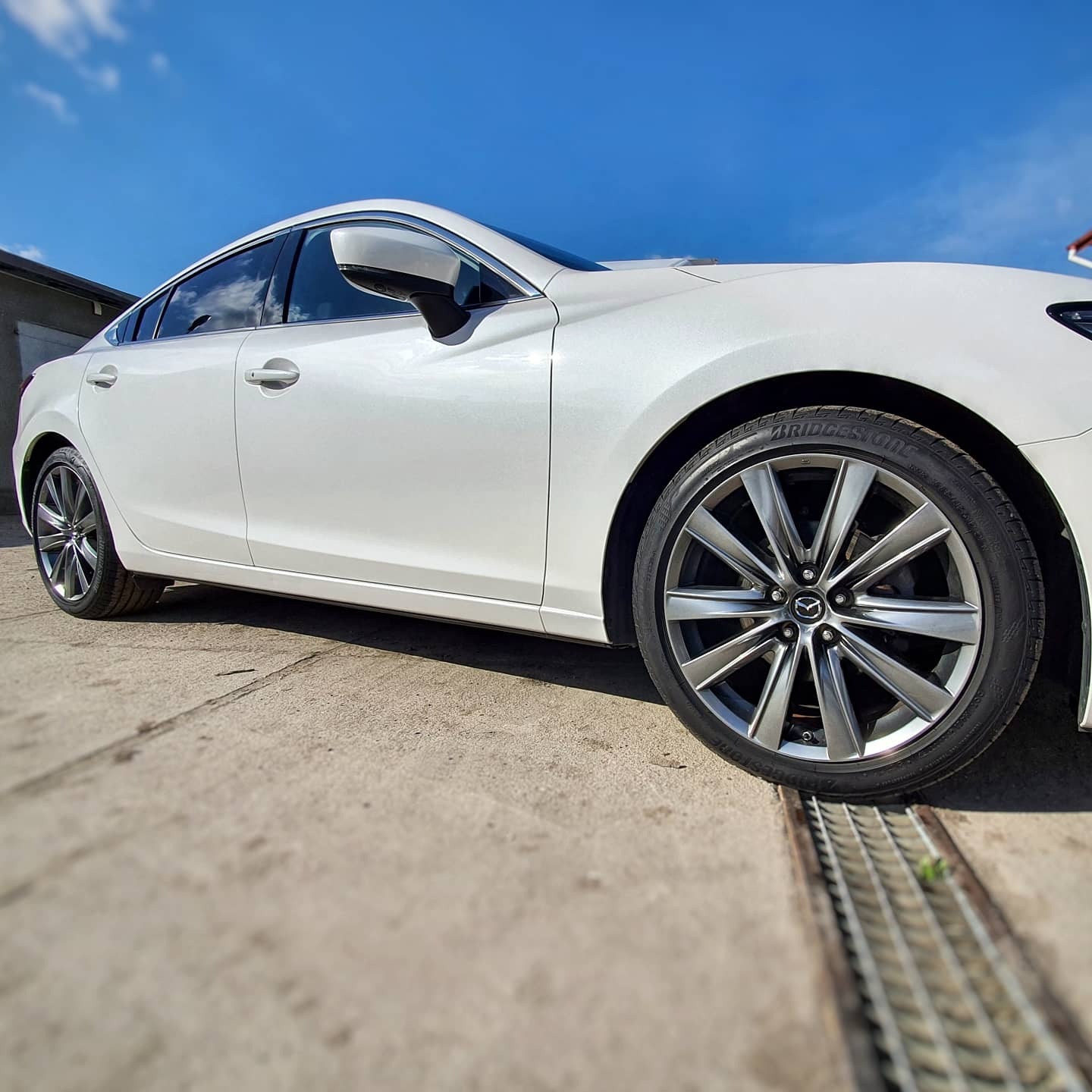 Biała Mazda 6 po umyciu, z widoczną oponą Bridgestone i felgą, sfotografowana z niskiej perspektywy na betonowym podłożu z kratką ściekową.