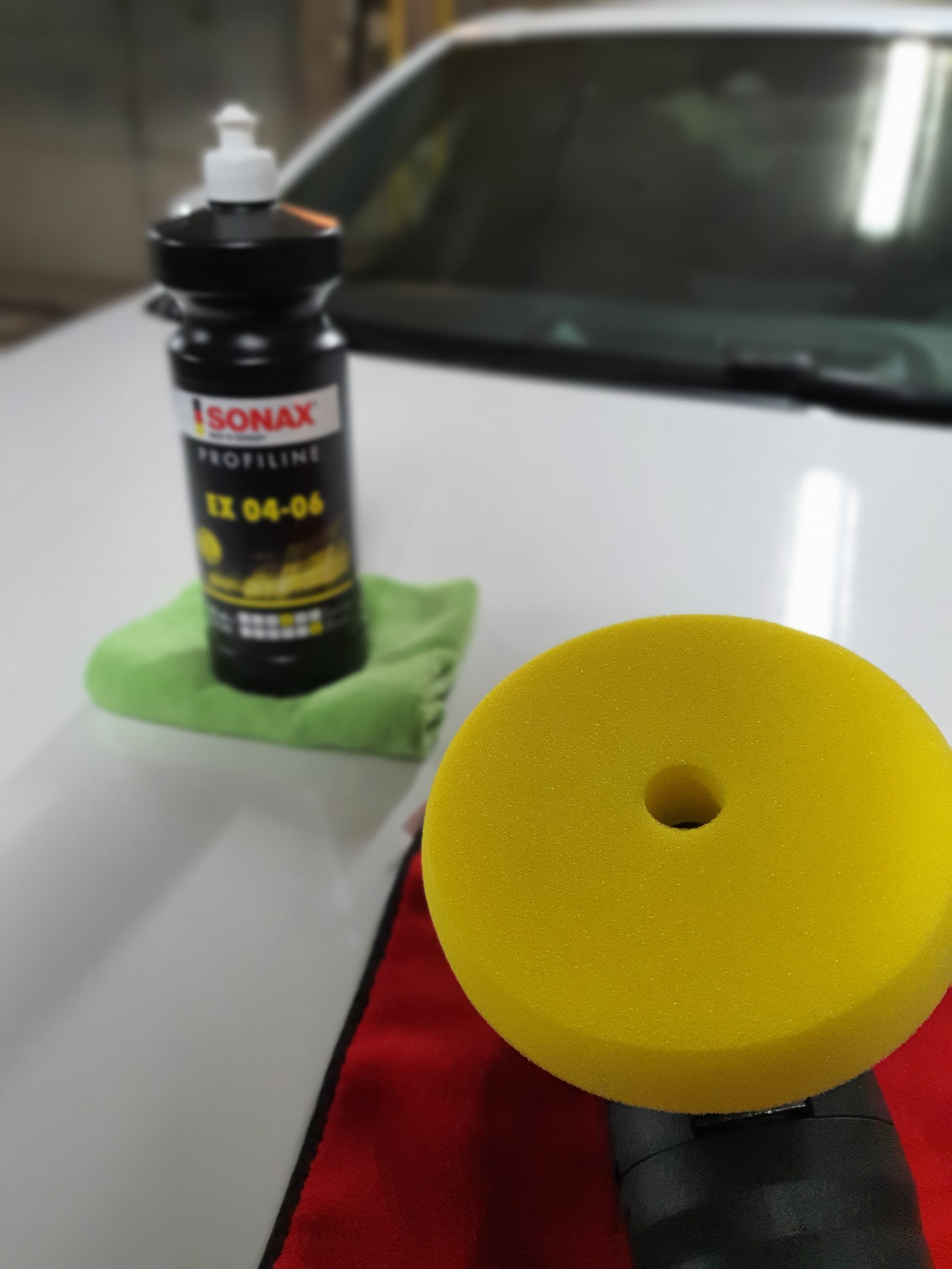 Butelka pasty polerskiej Sonax Profiline EX 04-06 na zielonej ściereczce, obok żółta gąbka polerska na czerwonej ściereczce, na tle białego samochodu.
