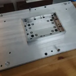 Aluminiowa płyta z frezowanym wgłębieniem i otworami, widoczne detale obróbki skrawaniem, pojemniki ze śrubami w tle.