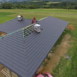 Ekipa dekarzy w czerwonych kombinezonach montuje ciemnoszarą blachodachówkę na nowym dachu domu jednorodzinnego, widok z lotu ptaka na tle zielonych pól i lasów.