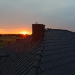 Widok z góry na ciemny dach z kominem w Jeżowem, na tle zachodzącego słońca.