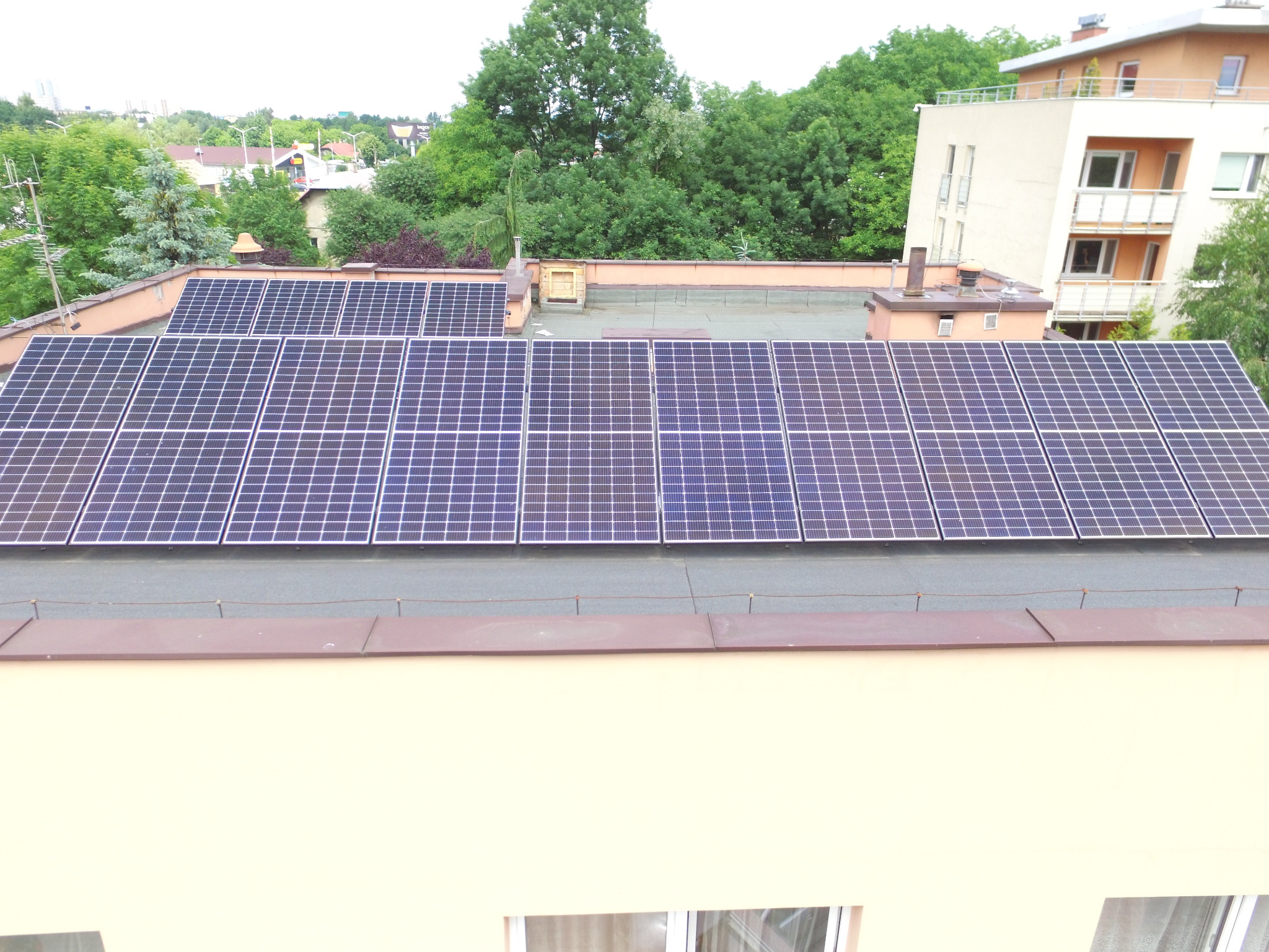 Panele fotowoltaiczne zamontowane na płaskim dachu budynku mieszkalnego, widok z góry, w tle zieleń drzew i zabudowa miejska.