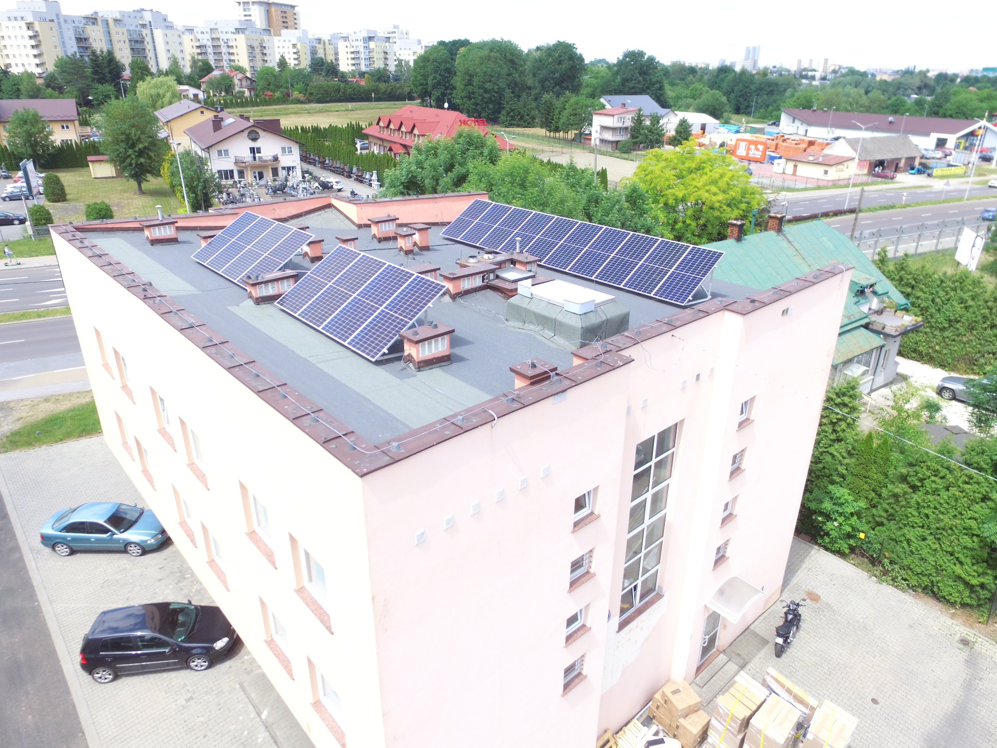 Widok z góry na budynek z zainstalowanymi panelami słonecznymi na płaskim dachu, na tle zabudowy miejskiej i zieleni.