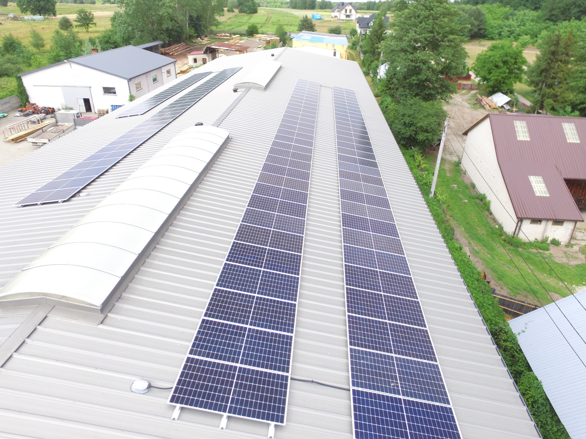 Widok z góry na zainstalowane panele słoneczne na falistym, metalowym dachu budynku przemysłowego z świetlikami dachowymi; w tle zabudowania gospodarcze i zieleń.