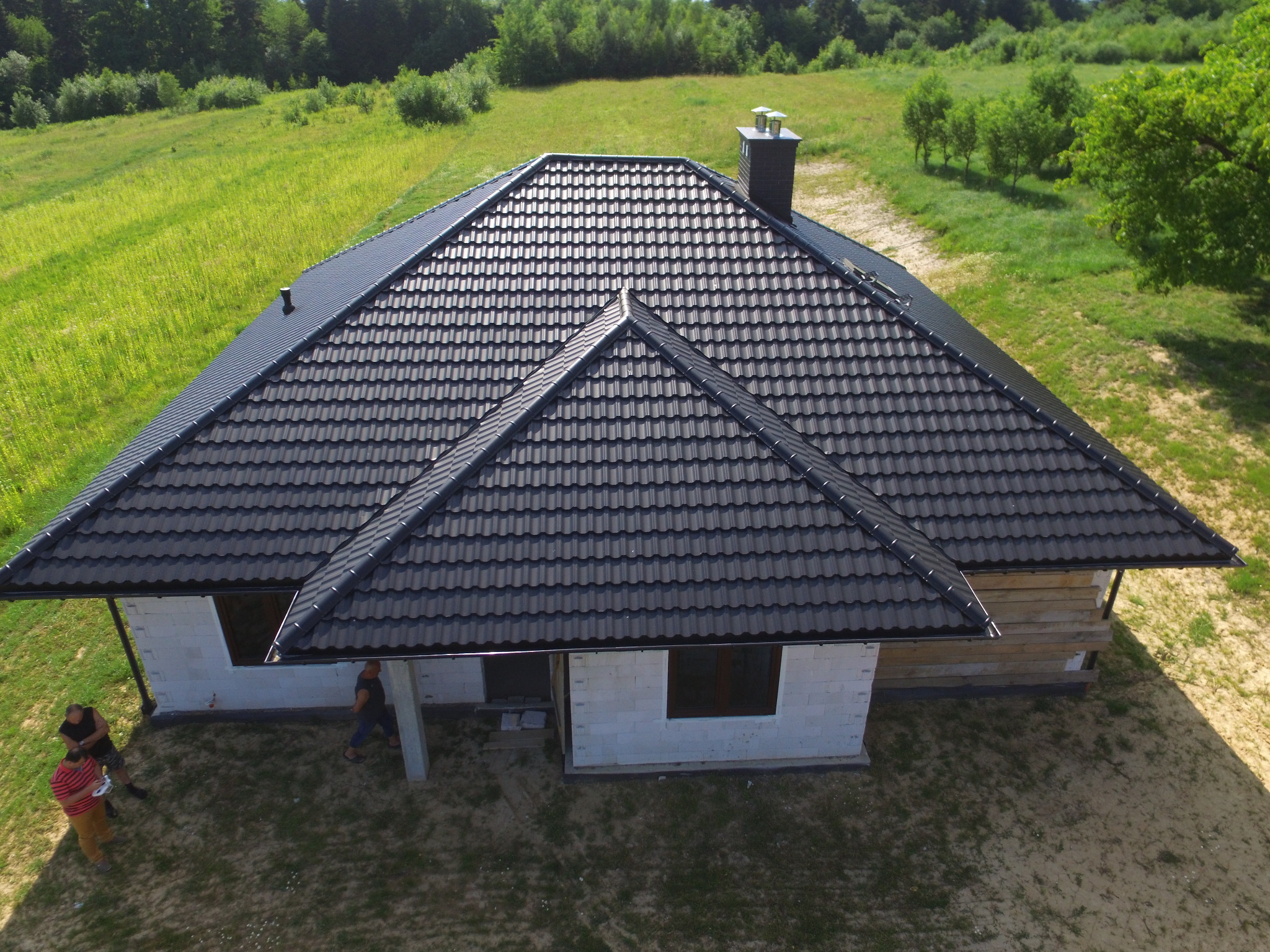 Widok z góry na dom w trakcie budowy z nowym, ciemnoszarym dachem z dachówki metalowej, komin z cegły, widoczni robotnicy i budynek w stanie surowym otwartym na tle zielonej łąki.