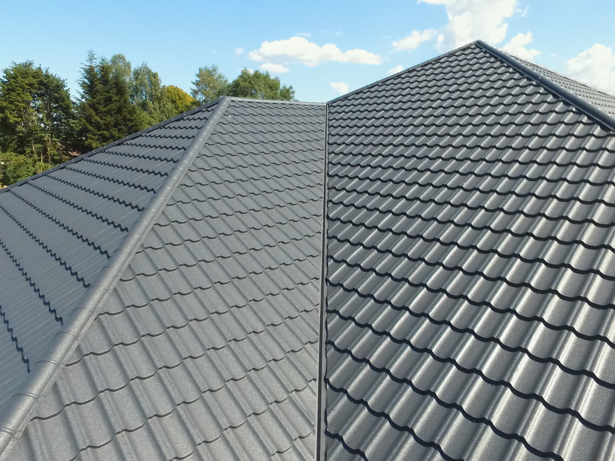 Szary dach z blachodachówki modułowej, widok z góry na kalenicę, w tle błękitne niebo i zieleń drzew.