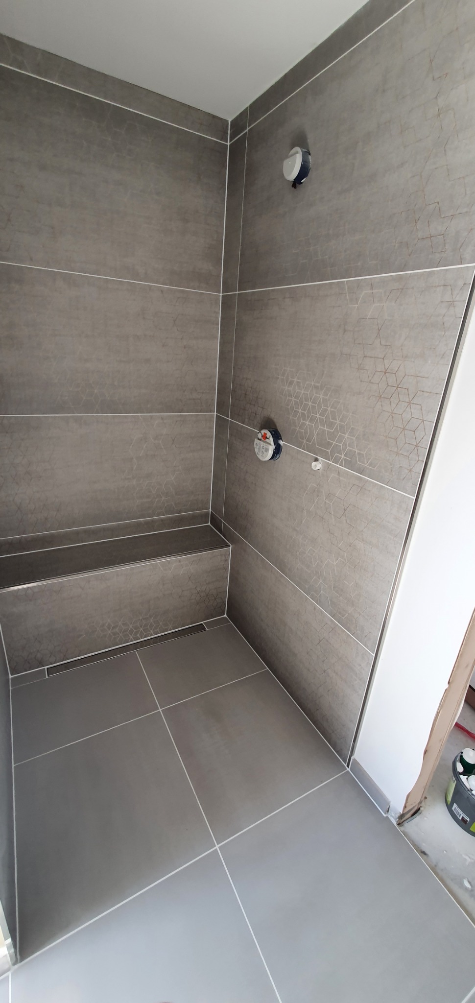 Szare, geometryczne płytki w kabinie prysznicowej z zamontowaną armaturą i odpływem liniowym w podłodze.