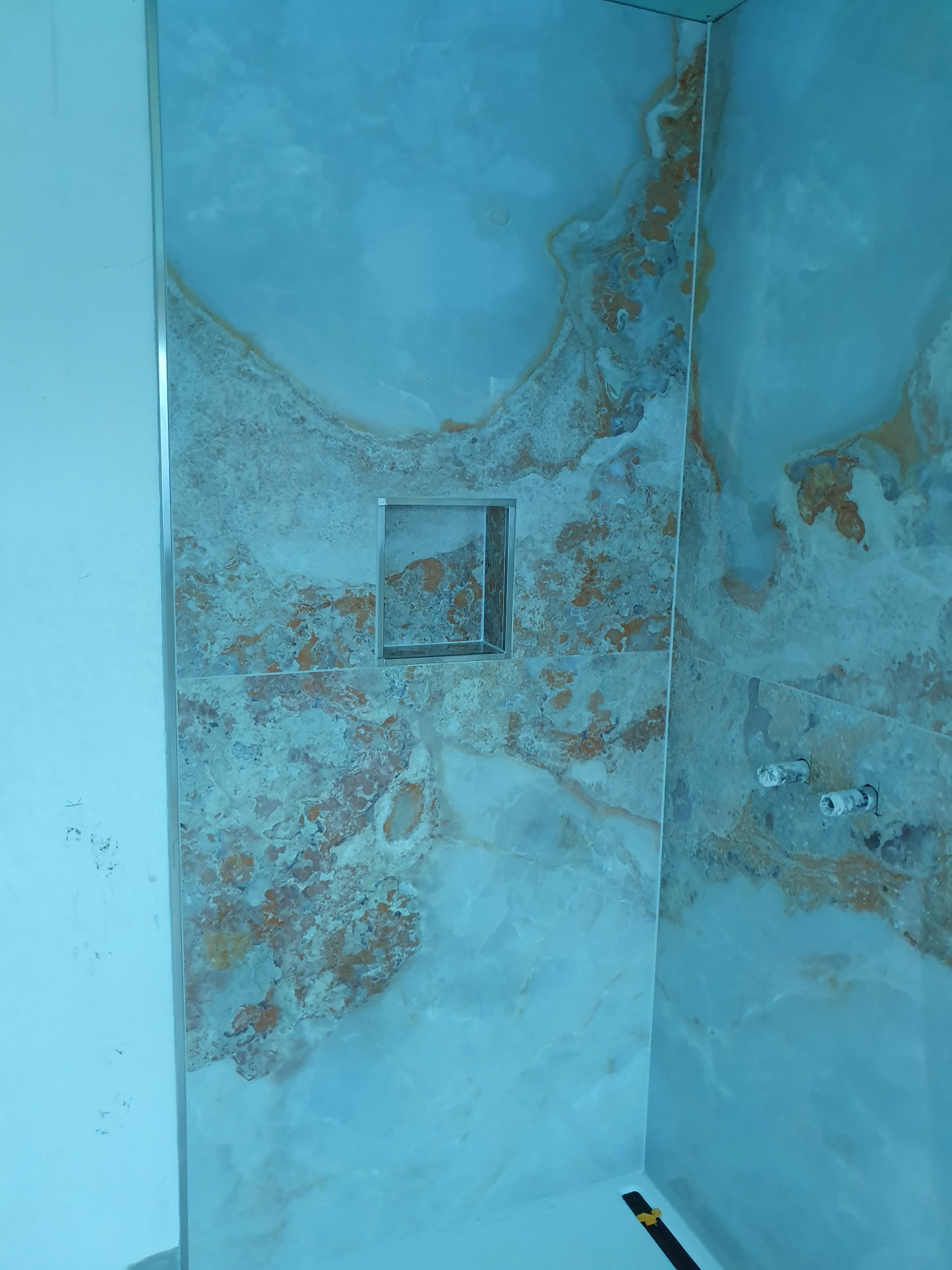 Wykończona ściana prysznicowa z płytek imitujących marmur w odcieniach błękitu i beżu, z wnęką na kosmetyki wykończoną metalową ramką i wystającymi rurami instalacji wodnej.