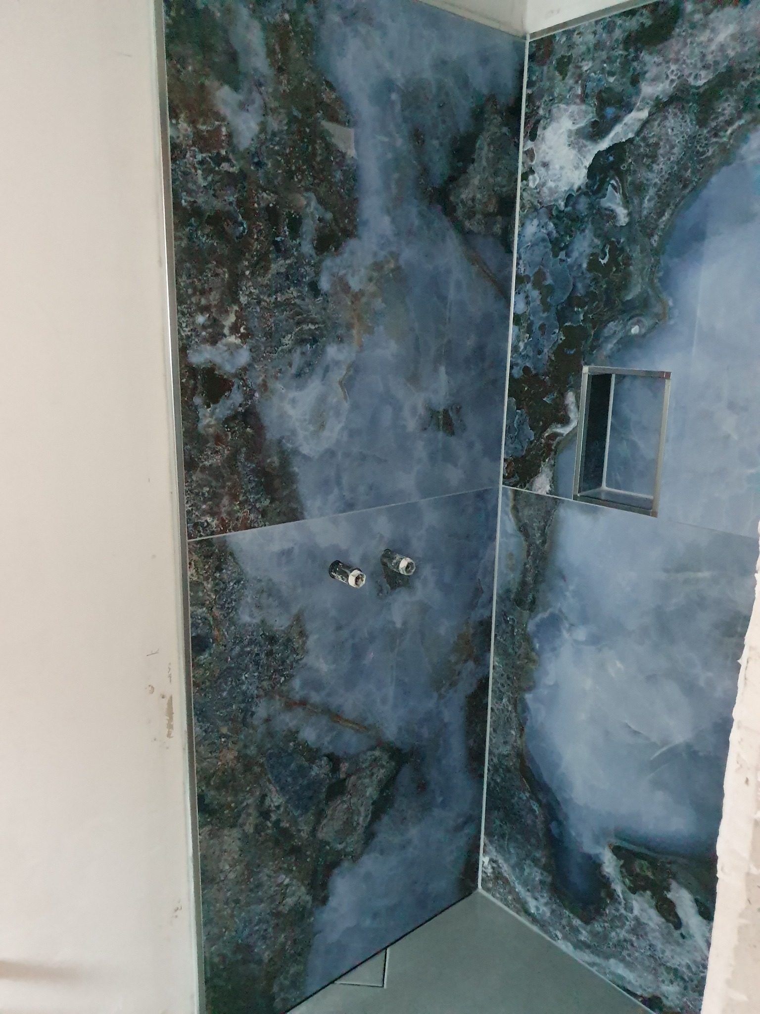 Ściana łazienki wyłożona płytkami imitującymi marmur w odcieniach błękitu i szarości, widoczne dwa zawory i wnęka prysznicowa z metalową ramką, fragment nieotynkowanej ściany.