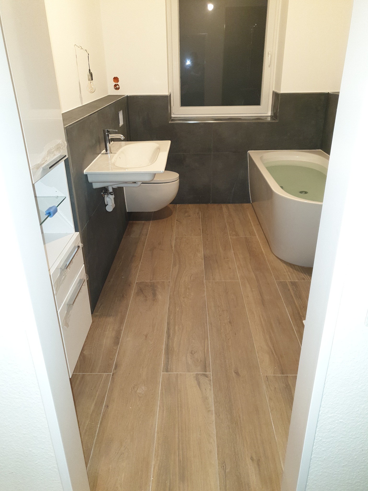 Wykończona łazienka z drewnopodobną podłogą, szarą glazurą na ścianach, białą wanną wypełnioną wodą, podwieszaną umywalką z toaletą oraz szafką z otwartymi półkami zabezpieczoną folią.