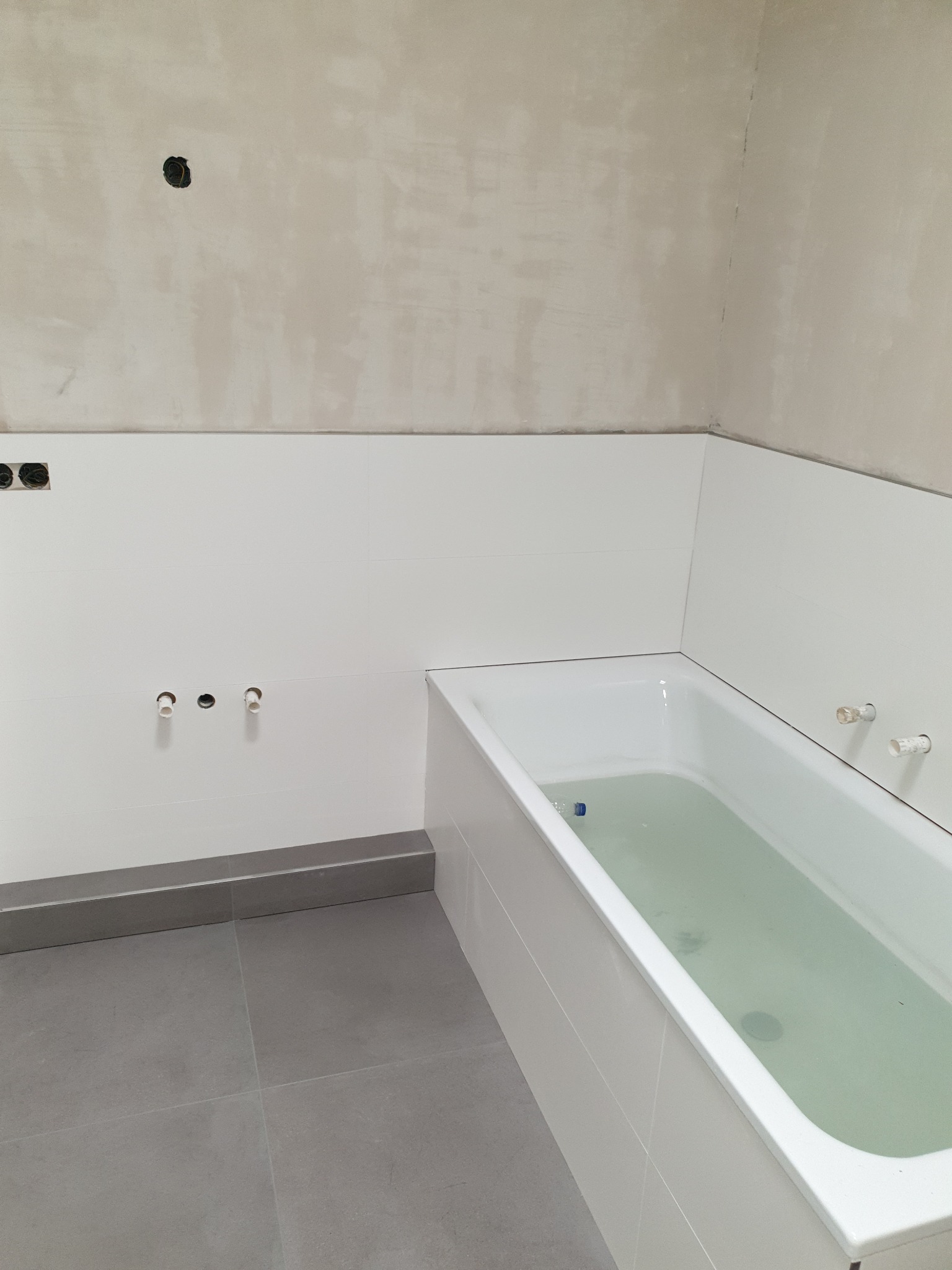 Wnętrze łazienki w trakcie remontu: białe płytki ścienne, szare płytki podłogowe, wanna wypełniona wodą, surowy tynk na górnej części ścian, widoczne instalacje hydrauliczne.