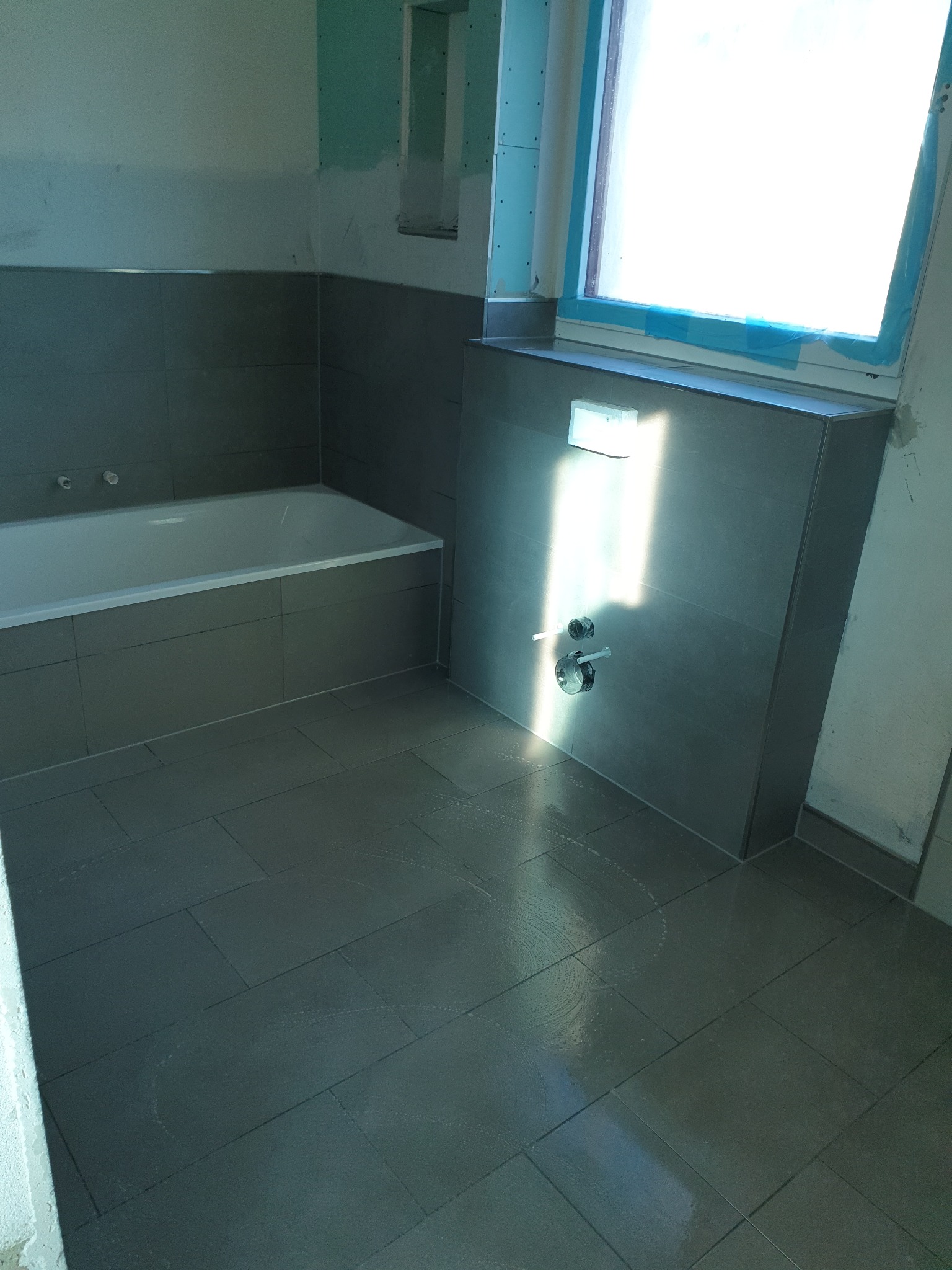 Szare płytki podłogowe i ścienne w łazience w trakcie remontu, widoczne ślady po fugowaniu, okno zabezpieczone folią, surowe ściany.