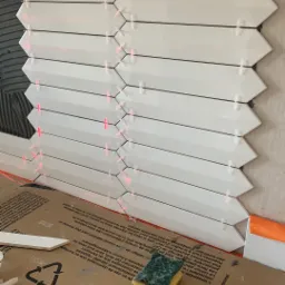 Układanie białych, podłużnych płytek o kształcie strzałki na ścianie z użyciem krzyżyków dystansowych i lasera wyznaczającego linię, widoczne narzędzia glazurnicze i opakowania.