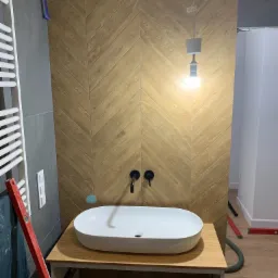 Łazienka w trakcie prac: umywalka nablatowa na drewnianym blacie, ściana wyłożona drewnopodobnymi płytkami ułożonymi w jodełkę, widoczny poziomica i niedokończona instalacja oświetleniowa.