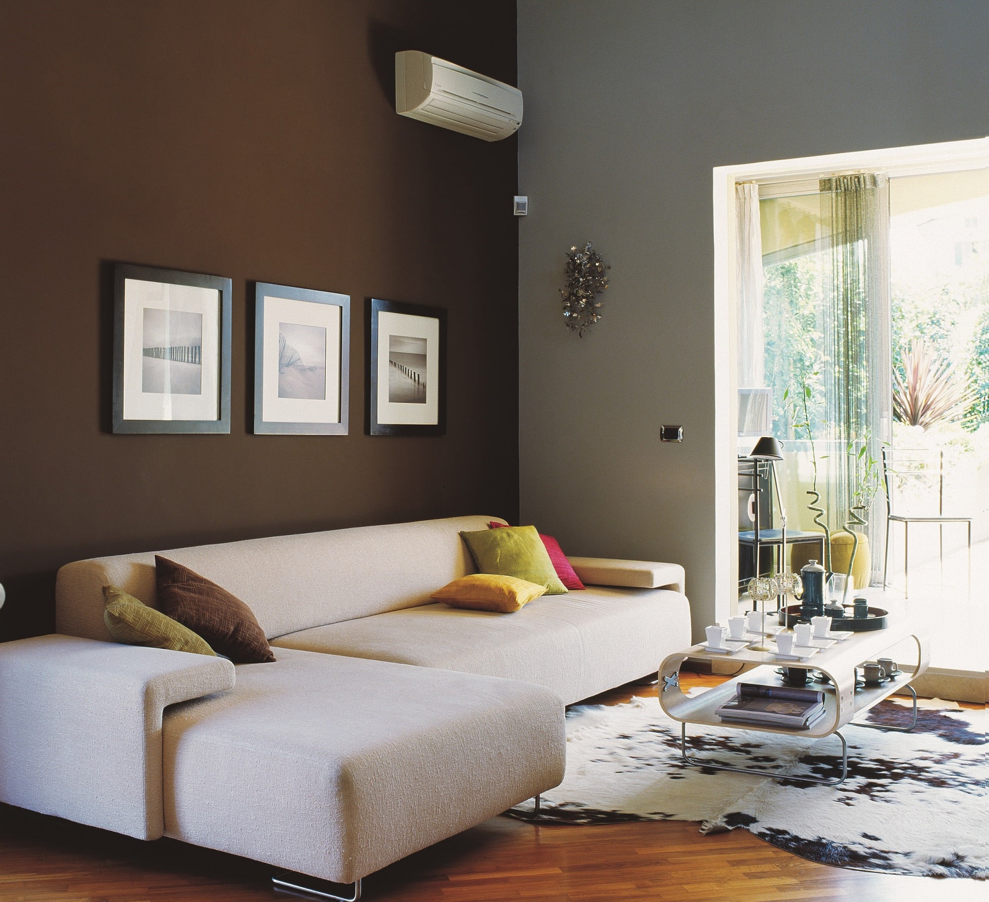 Nowoczesny salon z klimatyzacją ścienną, beżową sofą narożną, trzema obrazami na brązowej ścianie i dywanem w krowie łaty na parkiecie.