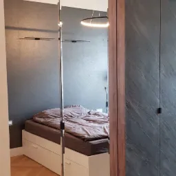 Szafa wnękowa z lustrzanymi drzwiami i ciemnymi panelami, odbicie sypialni z łóżkiem na podeście i parkietem w jodełkę.