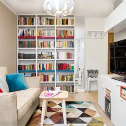 Jasny salon z dużą białą biblioteczką z kolorowymi akcentami, beżową sofą z poduszkami, stolikiem kawowym i dywanem w geometryczne wzory, telewizor na białej szafce RTV.